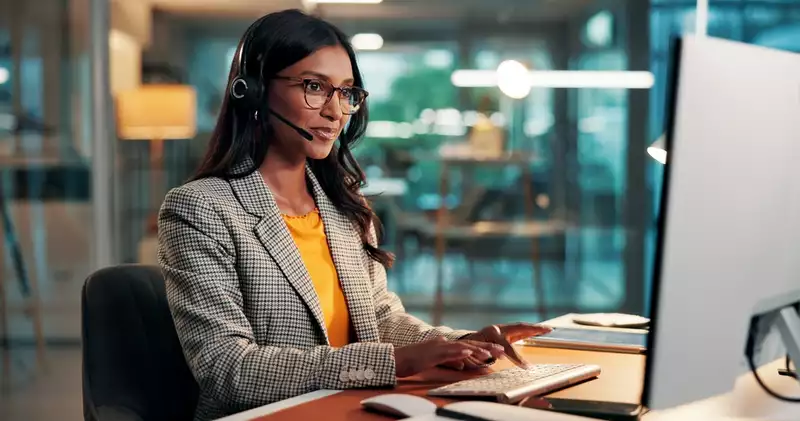 Business finance agent wearing a headset speaks with a prospect while working at a desktop computer.